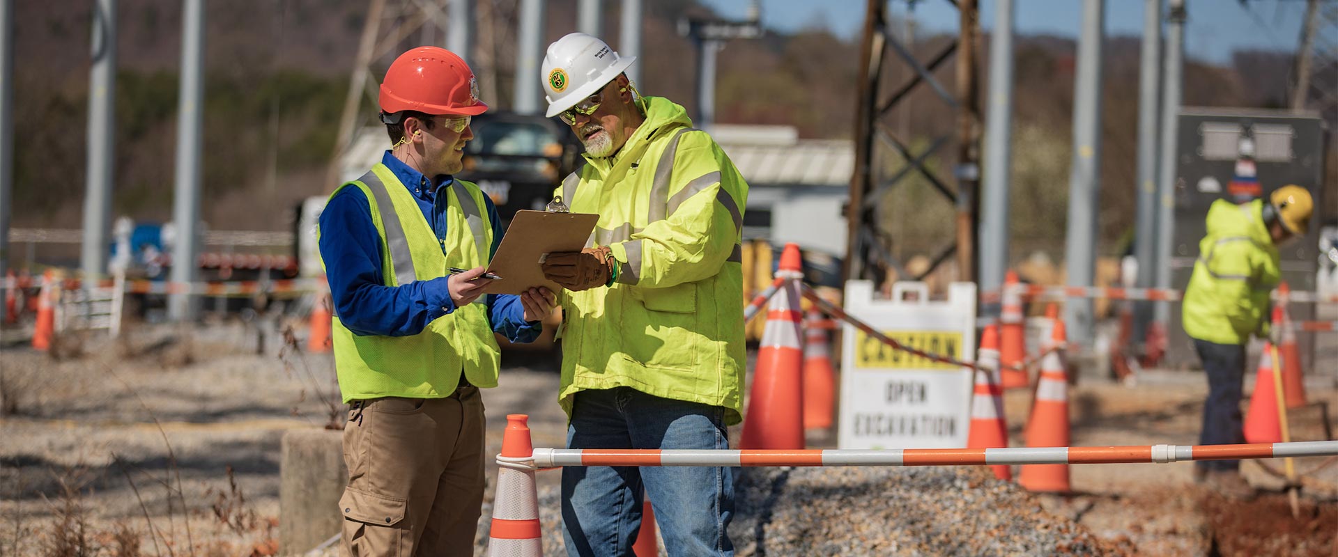 Two workers in yellow safety garments and hard hats stand at a work site and discuss something on a clipboard while another worker in yellow safety garb works in the background