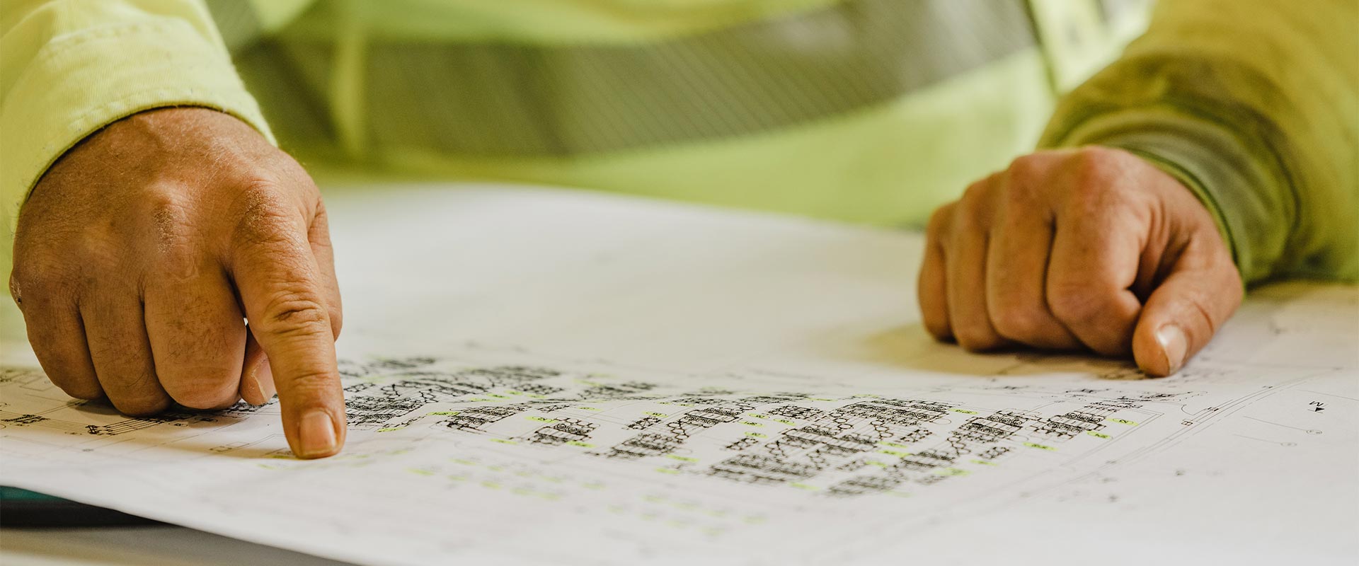 The hands of two workers resting on a paper set of plans; one hand is pointing at something on the page.