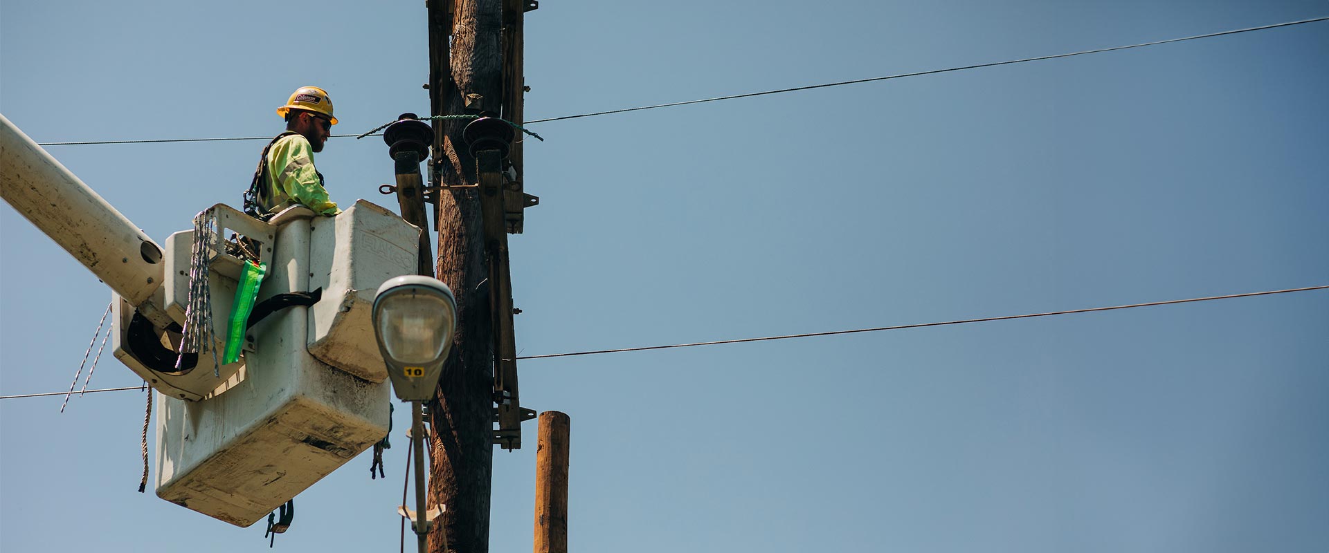 Man in yellow safety jacket and hard hat stands in the basket of an aerial lift and works on a utility pole beneath a clear blue sky.