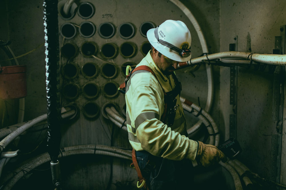 Man in yellow safety jacket and white hard hat stands in front of a series of circular holes in a concrete wall, some of which have thick electrical wires coming out of them.