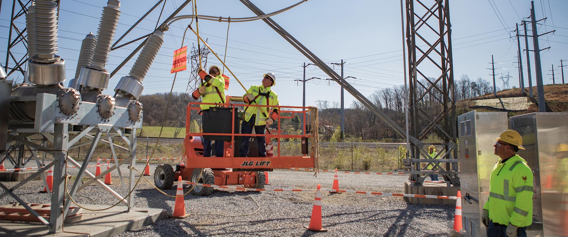 Two NRE workers in yellow safety jackets and hard hats stand on an orange mechanized lift and work on a large silver substation situated in a gravel lot while a two more NRE worker in yellow safety jacket and hard hat watches from the side and rear